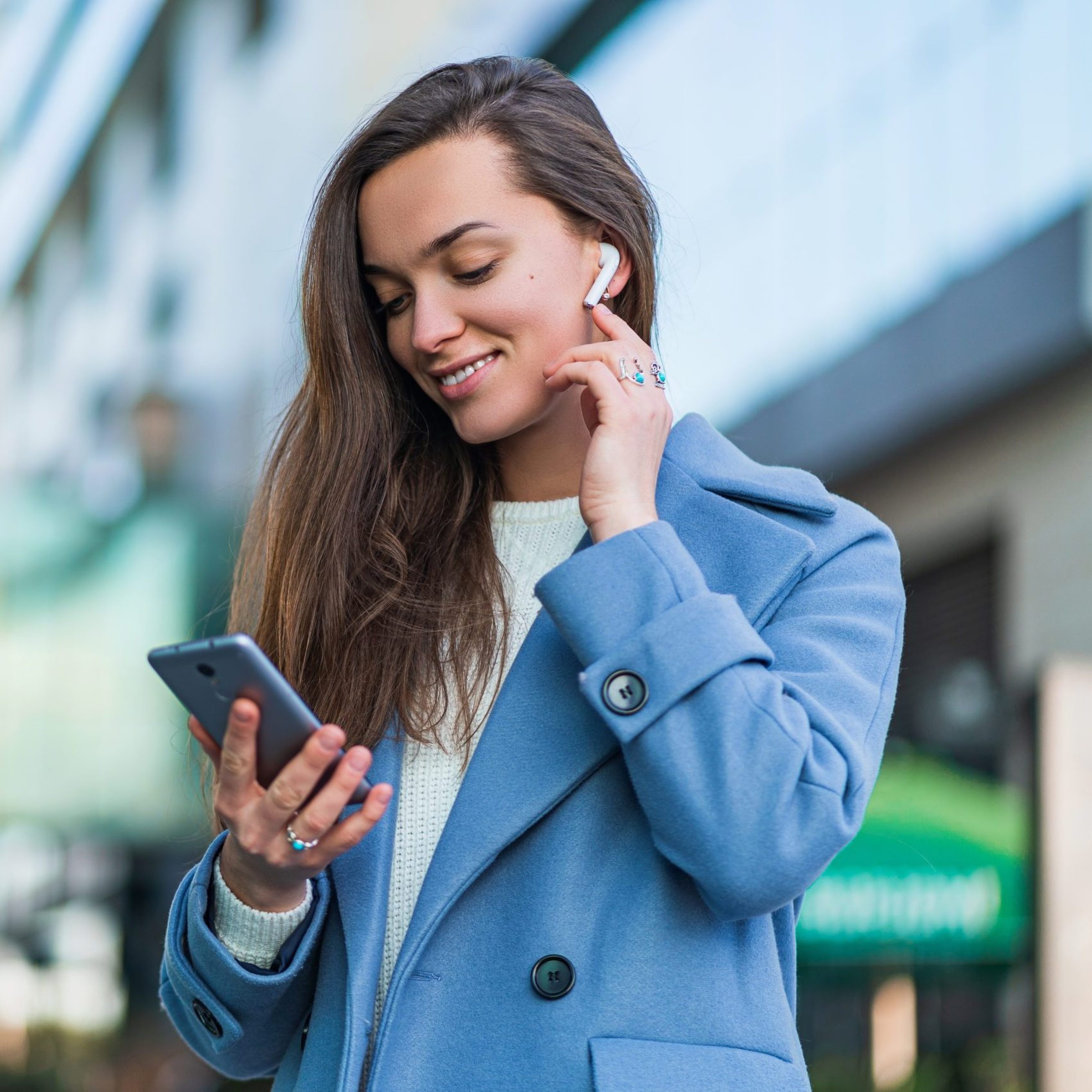 Stylish trendy happy joyful brunette woman in a blue coat holds a smartphone and using wireless white headphones for listen music in the city centre. Modern people